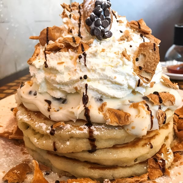 Good Eats' cannoli pancakes, topped with whipped cream, chocolate chips, and pastry pieces.