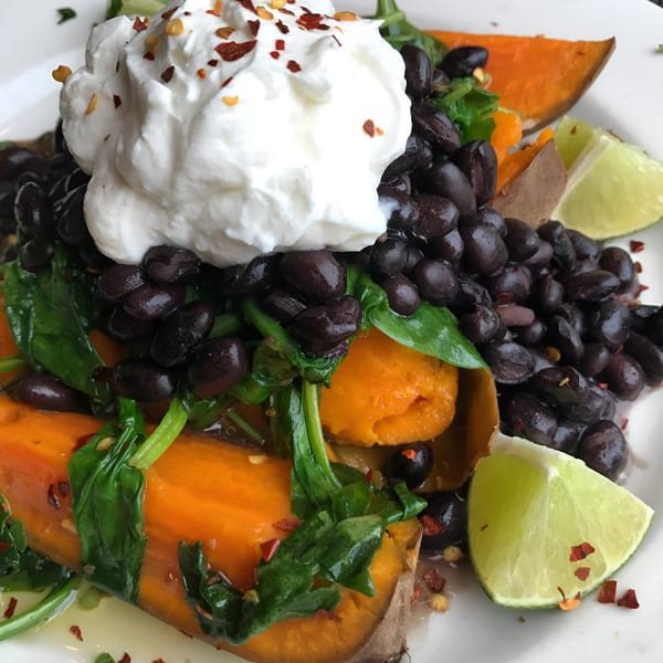 A baked sweet potato topped with black beans, spinach, sour cream, chili flakes, and lime wedges.