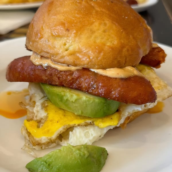 A Good Eats breakfast sandwich with sausage, avocado, fried egg, and sauce on a bun.