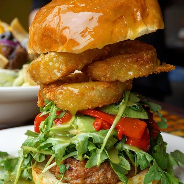 A Good Eats diner burger with onion rings, avocado, roasted red peppers, and arugula on a brioche bun.